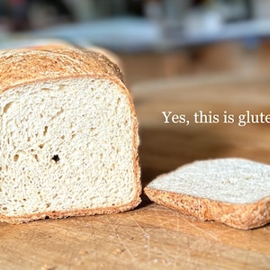 Puede incluir: Una hogaza de pan de color claro con corteza dorada, cortada para revelar una textura interior porosa. Junto a la hogaza hay una rebanada individual. El texto "Yes, this is gluten free" est&aacute; impreso en blanco a la derecha.