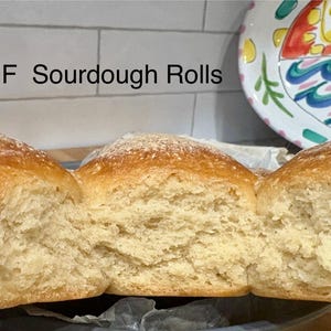 Puede incluir: Primer plano de panecillos de masa madre sin gluten. Los panecillos son de color marr&oacute;n dorado con una ligera capa de harina. El interior es de color beige claro y tiene una textura suave y aireada. Los panecillos se muestran sobre una superficie oscura.