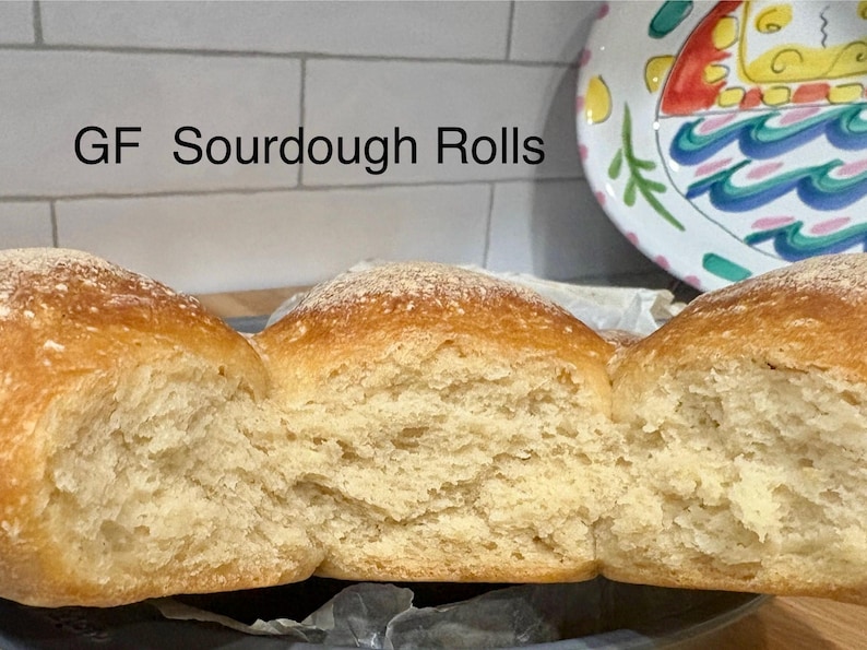 Puede incluir: Primer plano de panecillos de masa madre sin gluten. Los panecillos son de color marr&oacute;n dorado con una ligera capa de harina. El interior es de color beige claro y tiene una textura suave y aireada. Los panecillos se muestran sobre una superficie oscura.