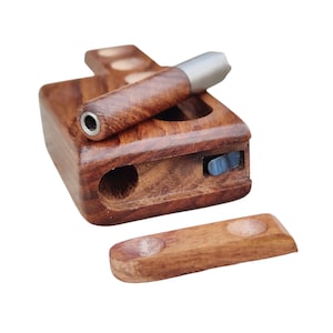 Puede incluir: Una caja de madera con una tapa deslizante, que contiene una pequeña pipa de madera con una punta de metal. La caja tiene un compartimento para guardar tabaco u otros materiales para fumar. La pipa está hecha de madera oscura y tiene un acabado natural.