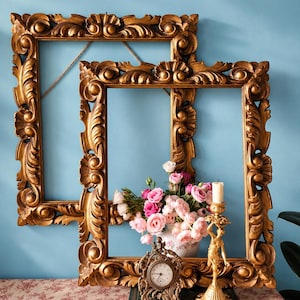 May include: Two rectangular gold picture frames featuring ornate carved scrollwork and floral motifs. In front, a small metal clock, a gold-toned figurine candle holder, and a bouquet of pink and white roses rest on a dark green book.