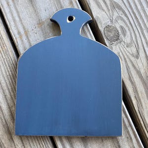 May include: A small, dark blue, wooden chopping board with a rounded top and a hole in the handle. The board is sitting on a wooden surface.