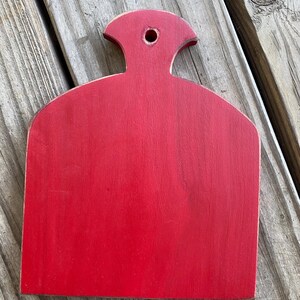 May include: A red wooden chopping board with a handle. The board has a rounded shape and a hole in the handle.