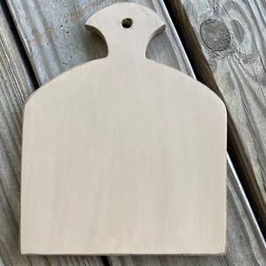 May include: A light brown wooden chopping board with a rounded top and a small handle. The board has a smooth, unfinished surface.