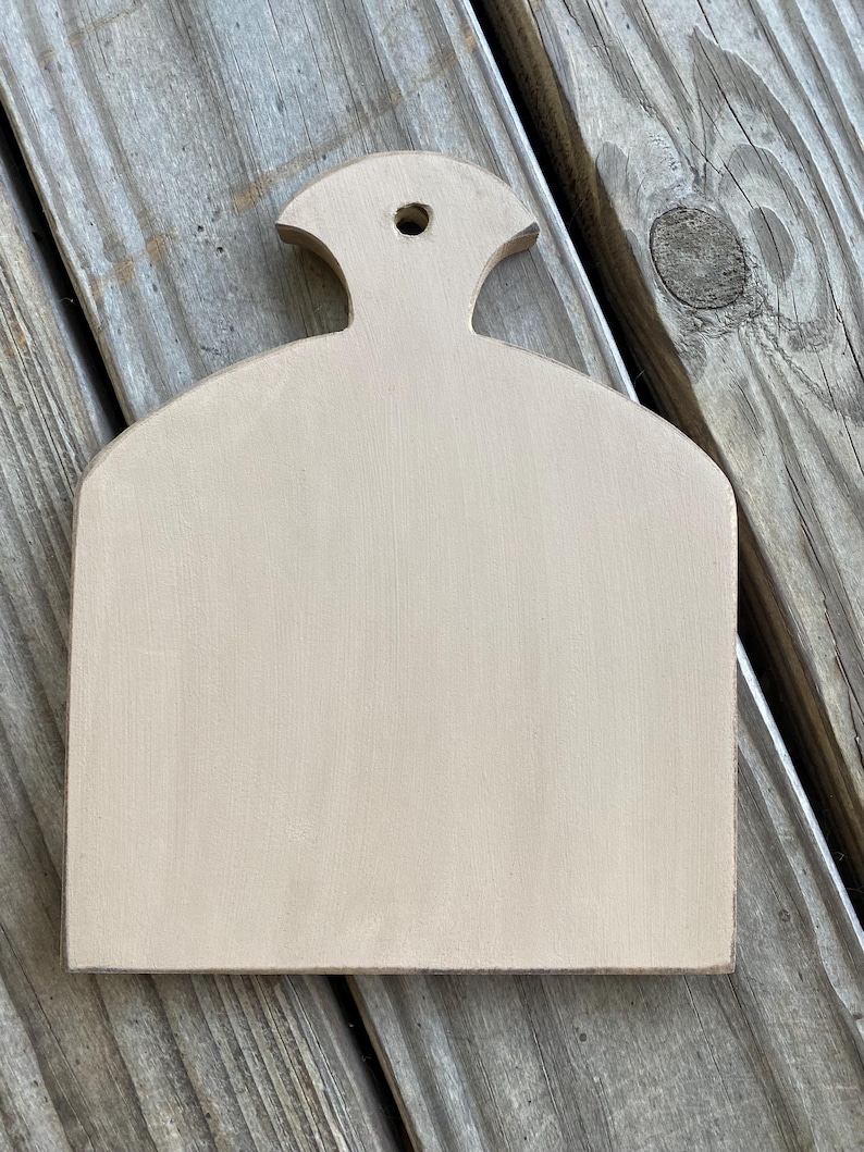 May include: A light brown wooden chopping board with a rounded top and a small handle. The board has a smooth, unfinished surface.