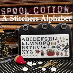 May include: A cross-stitch sampler with the alphabet and decorative elements, including a crow, scissors, and a strawberry pincushion. The text "A Stitchers Alphabet" is above the sampler. A vintage sewing machine and buttons are also visible.