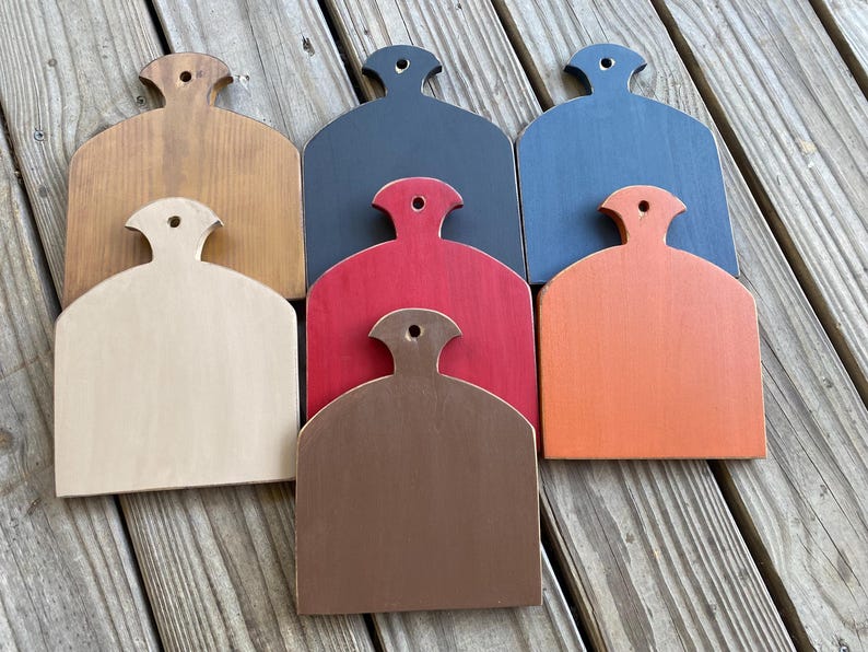 May include: Seven wooden chopping boards in various colours, including natural wood, red, orange, brown, black, and blue. The boards are all shaped like a rounded rectangle with a handle at the top.
