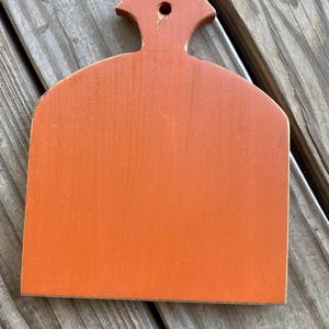 May include: Orange wooden chopping board with a handle in the shape of a rounded rectangle. The board is painted a bright orange colour.