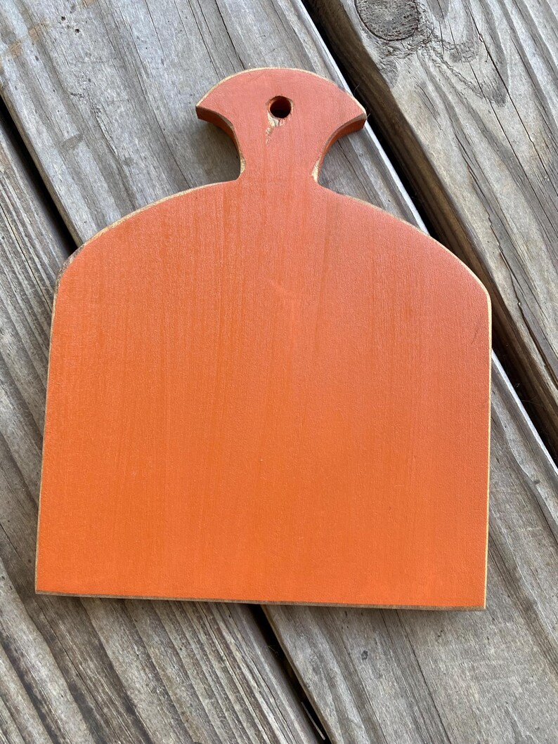 May include: Orange wooden chopping board with a handle in the shape of a rounded rectangle. The board is painted a bright orange colour.