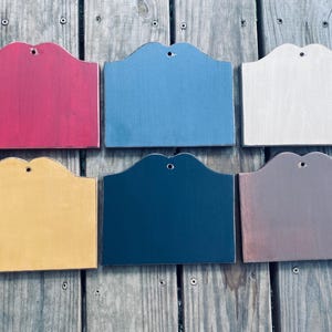 May include: Six wooden signs in various colors: red, blue, natural, yellow, navy, and brown. Each sign has a scalloped top edge and a small hole for hanging. The signs are arranged on a weathered wooden surface.
