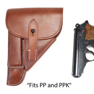 May include: A brown leather holster with a flap closure and a central strap, positioned between two semi-automatic pistols with dark metal frames and wooden grips. The text "Fits PP and PPK" is printed below the leather holster.