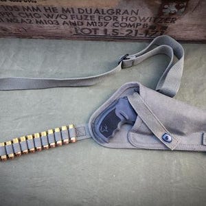 May include: A gray canvas holster with a strap, holding a black handgun. A matching gray belt is loaded with brass-tipped bullets. The background includes a wooden crate with text.