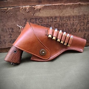 May include: A brown leather holster with a wooden handle and a row of bullets. The holster is secured with a metal button and is set against a wooden crate background.
