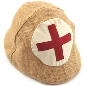 May include: Tan fabric cap with a white patch featuring a red cross emblem. The cap has a rounded shape and is made of a textured, woven material. The red cross is centered on the white patch.