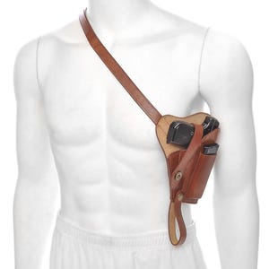May include: A brown leather shoulder holster with a strap, designed to be worn across the chest. The holster features a compartment for a handgun and an additional magazine pouch. The leather has a natural grain and a button closure.