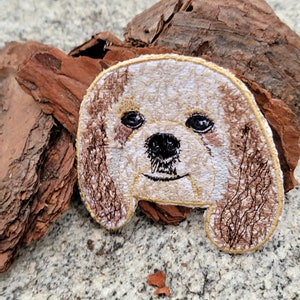 May include: Embroidered patch of a dog's face with brown fur and a white muzzle. The dog has brown eyes and a black nose.
