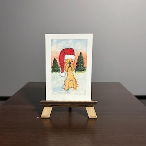 May include: Watercolor painting of a golden dog wearing a red Santa hat, set in a snowy winter scene with green trees and a colorful sky. The artwork is displayed on a small wooden easel.