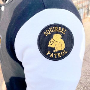May include: Black and white varsity jacket with a white patch on the sleeve that says "Squirrel Patrol" in yellow lettering and features a yellow squirrel graphic.