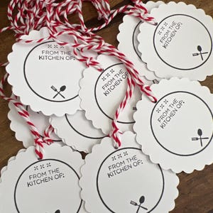 May include: White scalloped-edge gift tags with red and white twine. Each tag has a black circular border with the text "FROM THE KITCHEN OF:" and a spoon and fork icon. Ideal for homemade gifts.