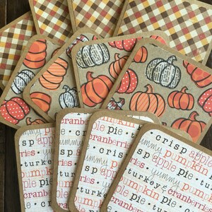 May include: A collection of square coasters with rounded corners. Some feature orange pumpkins on a tan background, others have a plaid pattern in fall colors, and some display text about Thanksgiving foods.