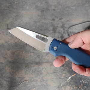 May include: A folding pocket knife with a silver-toned, flat-edged blade featuring a rectangular cutout near the spine. The handle is a solid matte blue with a visible pivot screw and a contoured grip design, held in a horizontal position.