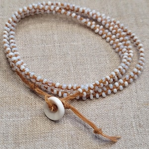 May include: A wrap bracelet featuring rows of small, white beads strung on a brown cord. The bracelet is secured with a white, circular button and a knotted cord closure. The design is simple and elegant, suitable for everyday wear.