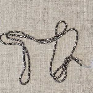 May include: A strand of silver-toned beads, arranged in a loose, looped shape on a textured beige fabric background.