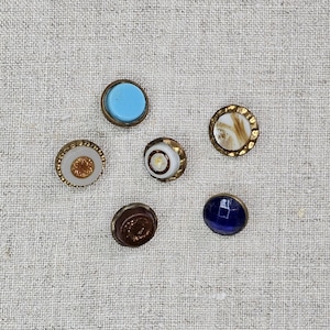 May include: A collection of six vintage buttons with various designs and colors. The buttons feature a range of colors, including blue, brown, white, and gold, with intricate detailing and metal rims. The buttons are arranged on a textured, light-colored fabric.