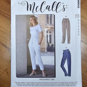 May include: McCall's sewing pattern M8045 featuring a woman in white pants and a white t-shirt. The pattern includes multiple view options. The size is E5 (14-16-18-20-22). The hashtag is #AbbieMcCalls.