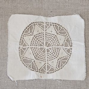 May include: A white cotton square with a delicate, intricate lace design in the center. The lace is a circular pattern with a starburst design.