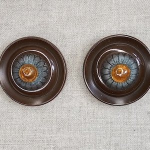 Peut inclure: Deux plats en céramique brune avec un motif floral bleu et jaune. Les plats ont un bord surélevé et un centre légèrement concave.