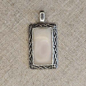 May include: A rectangular pendant with a light-colored, possibly agate, center stone. The stone is framed by a silver-colored metal with a decorative, woven-like pattern. The pendant has a small loop at the top for a chain.