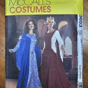 May include: McCall's Costumes sewing pattern 3653, size AA (6-8-10-12). The envelope features two floor-length gowns: one in blue with a patterned front panel and wide sleeves, and another in brown with white sleeves and side lacing details.