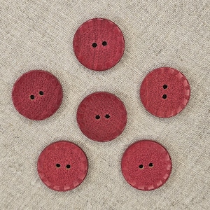 May include: A collection of six round, burgundy-colored buttons with two holes each. The buttons have a textured surface and are arranged on a neutral, textured fabric background. These buttons are suitable for sewing and craft projects.