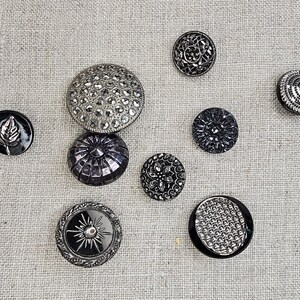 May include: Assortment of eight black and silver buttons with various designs. The buttons are round and have a raised design.