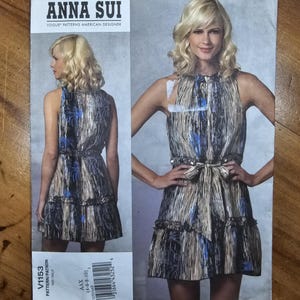 May include: An Anna Sui Vogue Patterns American Designer sewing pattern envelope, model V1153, featuring a sleeveless dress with a high neckline, a tie-waist, and a ruffled hem in a metallic gold, black, and blue abstract print on the front cover.