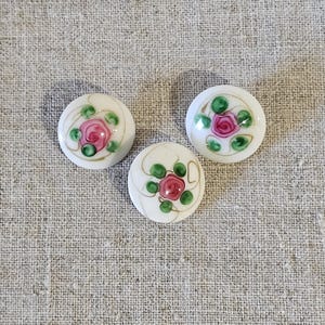 Peut inclure: Trois boutons en céramique blanche avec un motif de rose rose et de feuilles vertes. Les boutons ont une bordure dorée.