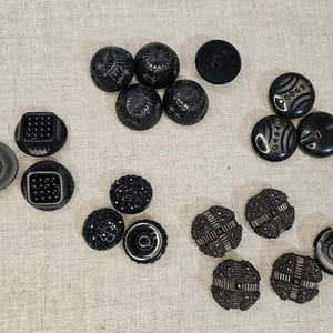 May include: A collection of various black buttons with different designs. Some buttons are round with textured surfaces, while others are square or have decorative patterns. The buttons are arranged on a neutral-colored fabric.