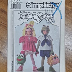 May include: Simplicity sewing pattern featuring Jim Henson's Muppet Babies. The pattern includes instructions for Miss Piggy and Kermit costumes. The package is white with a colorful illustration of the characters. The pattern is size A.