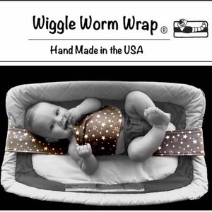 May include: A baby wearing a brown and white star patterned wrap is lying in a grey and white cot. The wrap is labelled "Wiggle Worm Wrap" and "Hand Made in the USA".
