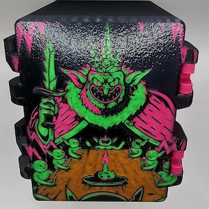 May include: Black box with a neon green and pink goblin illustration. The goblin, holding a sword, is surrounded by smaller goblins around a table with a candle. The box has pink accents.