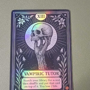 May include: A holographic card with a black border features a skull held by a skeletal hand. The card reads "VAMPIRIC TUTOR" with instructions below. Floral and geometric designs frame the central image. The card has the Roman numeral XIII at the top.
