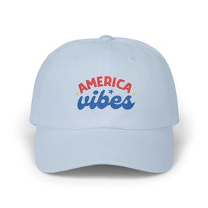 May include: A light blue baseball cap featuring the embroidered text AMERICA in red block letters and vibes in blue cursive script. A small blue star is positioned between the words, with two tiny yellow stars flanking the text design.