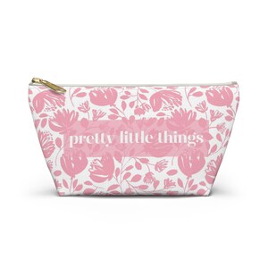 May include: A white cosmetic bag with a pink floral pattern and the text "pretty little things" in the center. The bag has a gold zipper and a rectangular shape.