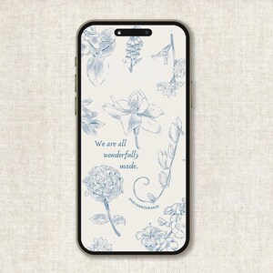 May include: A smartphone displays a floral design phone case. The case features blue botanical illustrations of various flowers and foliage against a cream background. The text "We are all wonderfully made" is visible, along with the artist's signature.