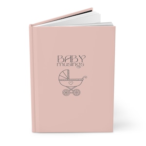 May include: A blush pink journal with the words "BABY musings" and a baby carriage illustration. The journal is open, revealing white pages. The cover is a solid, soft pink color.