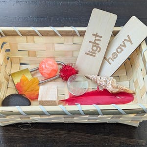 May include: A woven basket filled with various objects for a sensory activity. Includes wooden labels that read "light" and "heavy", a red feather, a seashell, a rock, and other small items.