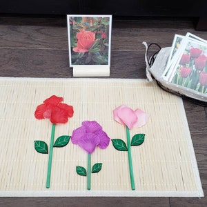 May include: Three felt roses on a bamboo mat. The roses are red, pink, and purple. There is a picture of a pink rose in a frame and a basket of seed packets in the background.