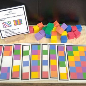 Puede incluir: Un conjunto de cubos de espuma de colores y tarjetas con instrucciones para un juego de emparejamiento de cubos. Las tarjetas muestran diferentes patrones de cuadrados de colores. Las instrucciones sugieren construir una torre, hacer una ciudad de cubos y tener un concurso de apilamiento de cubos.