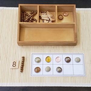 May include: A wooden tray with compartments containing wooden beads, number tiles, and small glass stones. A wooden number tile with the number 8 is on a bamboo mat. A white card with a grid of nine squares contains eight glass stones of different colors.